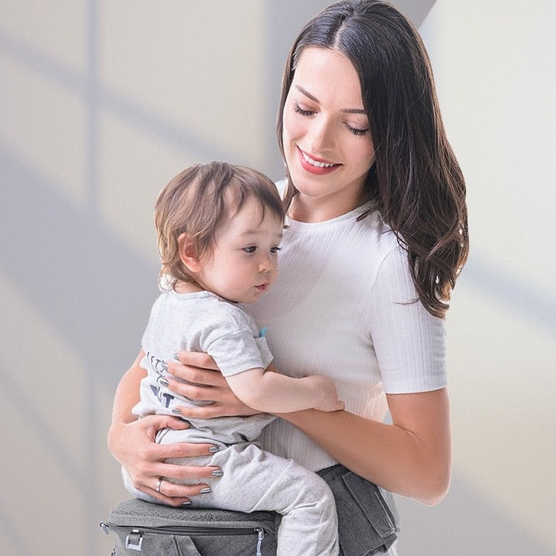 a happy young woman is holding her baby with ease as her little one is sitting on the baby carrier